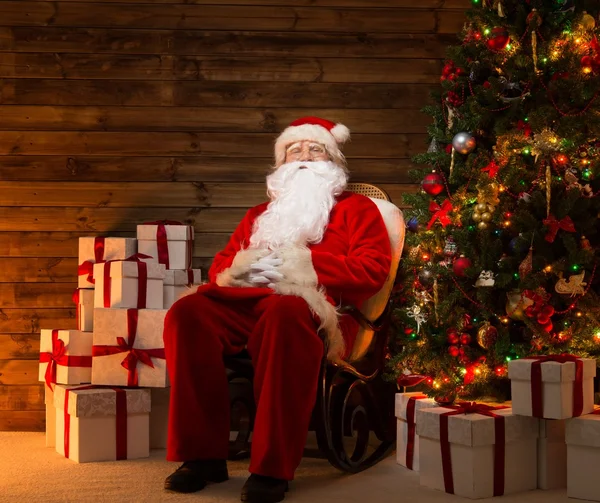 Papai Noel sentado em cadeira de balanço em interior de casa de madeira com caixas de presente ao seu redor — Fotografia de Stock