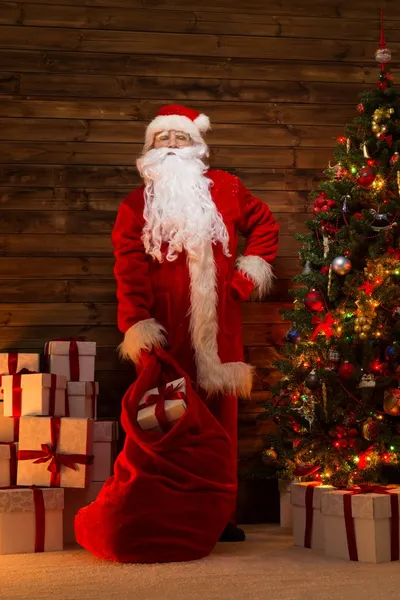 Papai Noel em casa de madeira interior com saco cheio de presentes de Natal — Fotografia de Stock