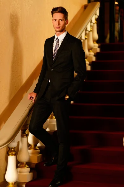 Jeune bel homme en costume noir debout sur les marches de l'intérieur de luxe — Photo