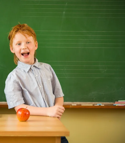 Pequeno estudante ruivo engraçado perto de quadro negro com maçã em uma mesa — Fotografia de Stock