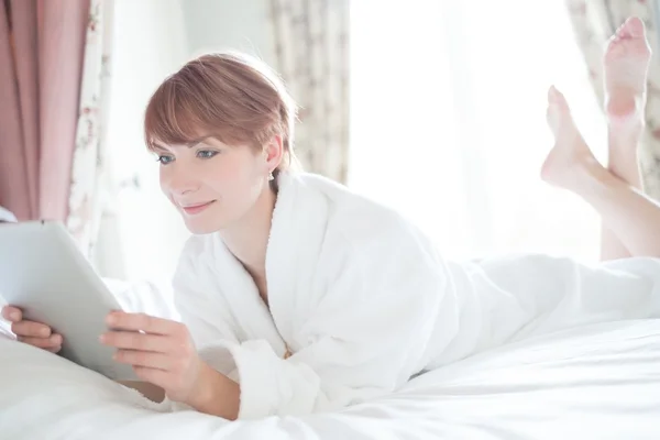 Schöne Frau im Bademantel auf einem Bett mit Tablet-PC liegend — Stockfoto