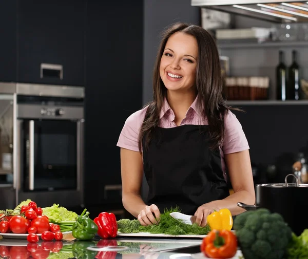 Glad ung kvinna i förkläde på modernt kök skära grönsaker — Stockfoto