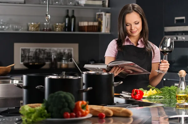 Vrolijke jonge vrouw in schort op moderne keuken met kookboek en glas wijn — Stockfoto