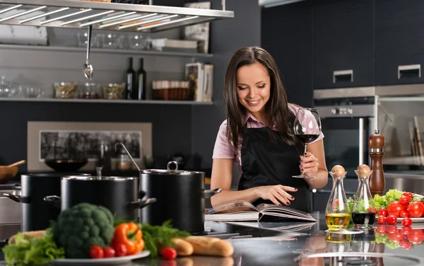 Joyeux jeune femme dans tablier sur cuisine moderne avec livre de cuisine et verre de vin — Photo