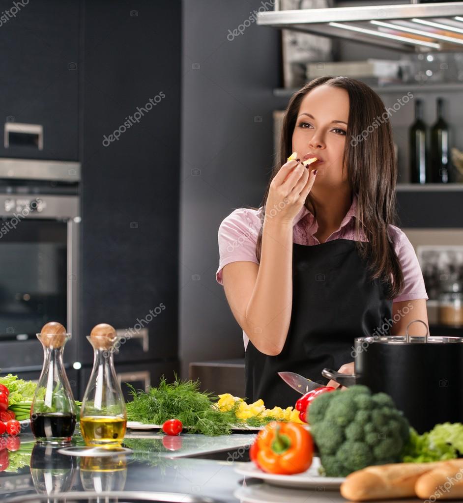 Glückliche junge frau in schürze auf moderne küche essen zu kosten beim kochen stockdatei