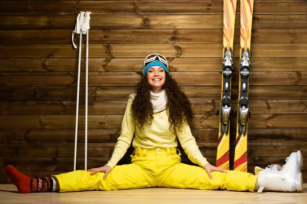 Glad kvinna med skidor och ski stövlar sitter på ett golv nära trävägg — Stockfoto