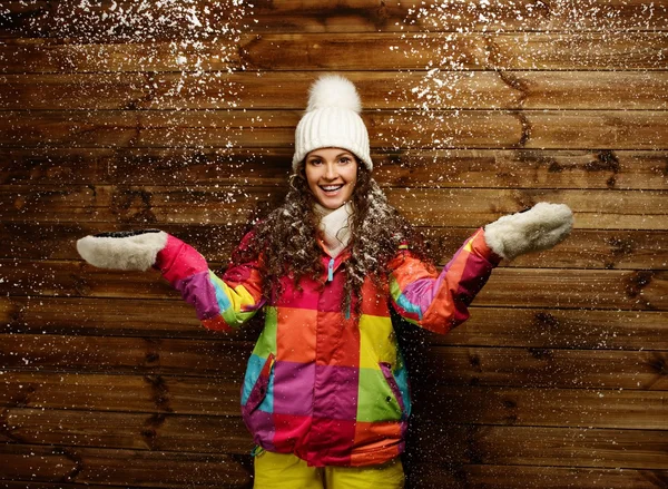 Mujer sonriente en chaqueta de esquí y sombrero blanco de pie contra la pared de la casa de madera bajo la nieve —  Fotos de Stock