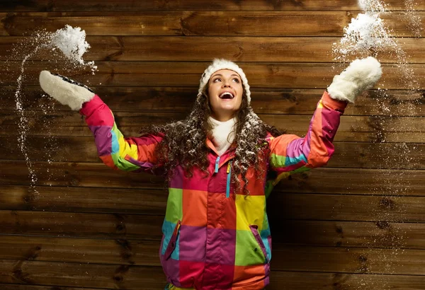 Mujer sonriente en chaqueta de esquí y sombrero blanco de pie contra la pared de la casa de madera bajo la nieve —  Fotos de Stock