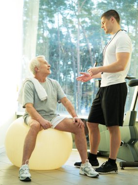 Personal trainer explains to a senior man how to do exercise on a fitness ball clipart