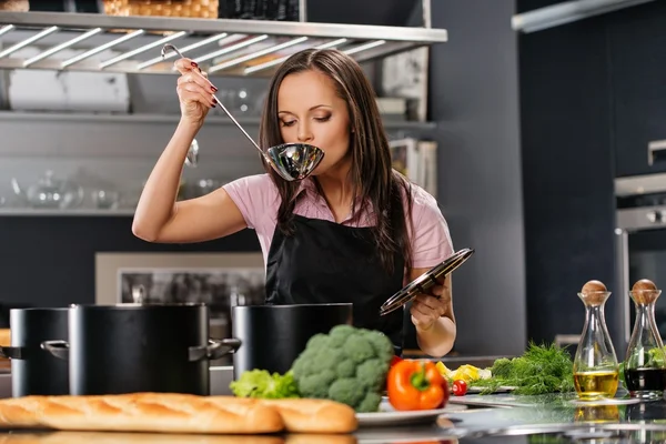 Joyeux jeune femme dans tablier sur cuisine moderne avec dégustation de louche de pot — Photo