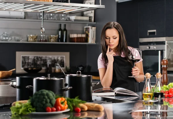 Cheerful young woman in apron on modern kitchen with cookbook and glass of wine — Stock Photo, Image