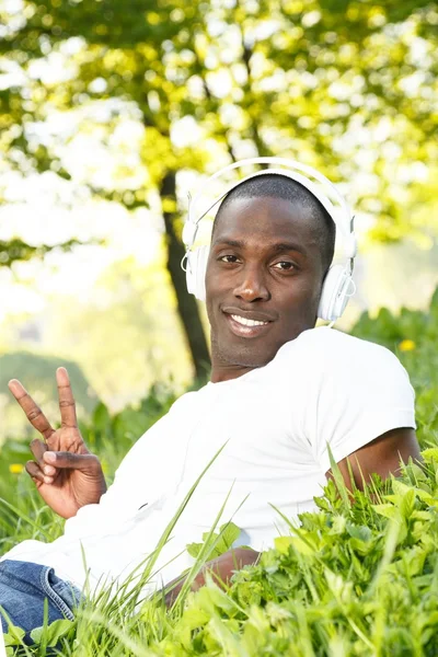 Positive young african american man in white shirt listens music in a park — Stock Photo, Image