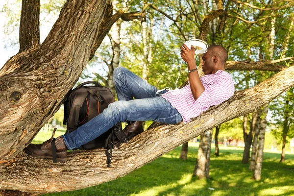 Jonge Afrikaanse Amerikaan liggend op een boomtak in een park en luistert naar muziek — Stockfoto