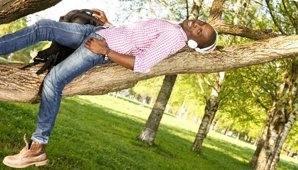 Young african american lying on a tree branch in a park and listens to music — Stock Photo, Image