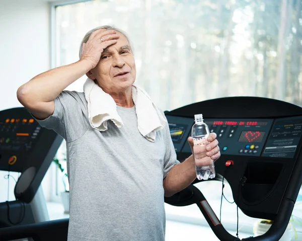 Tired senior man on a treadmill with towel and bottle of water — Stock Photo, Image