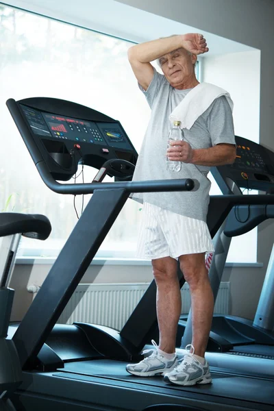 Tired senior man on a treadmill with towel and bottle of water — Stock Photo, Image