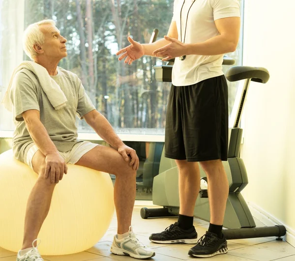 Entrenador personal explica a un hombre mayor cómo hacer ejercicio en una pelota de fitness — Foto de Stock