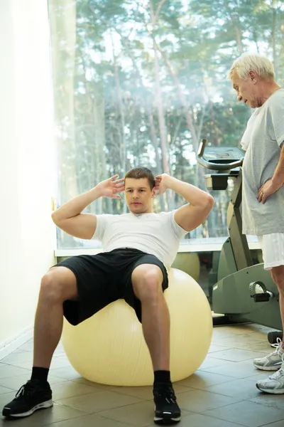 Treinador pessoal mostra a um homem sênior como fazer exercício em uma bola de fitness — Fotografia de Stock