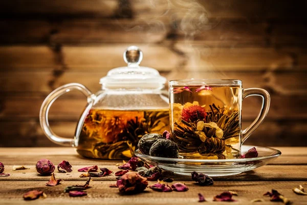 Teapot and glass cup with blooming tea flower inside against wooden background — Stock Photo, Image