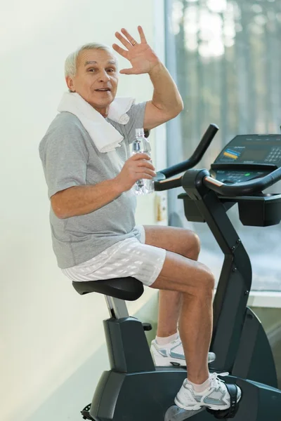 Uomo anziano con bottiglia d'acqua in bicicletta in un fitness club — Foto Stock