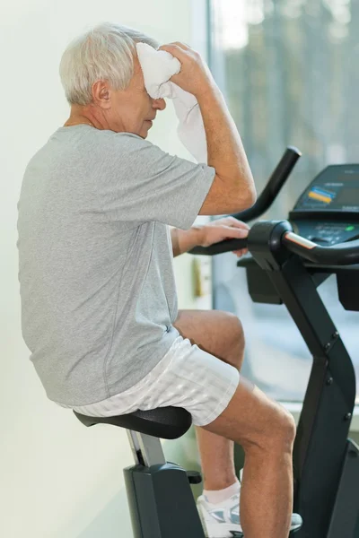 Tired senior man with towel on exercise bike in fitness club — Stock Photo, Image