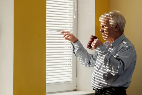 Senior mit Tasse blickt durch Jalousie aus dem Fenster — Stockfoto