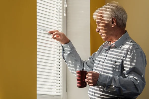 Uomo anziano con coppa che guarda fuori dalla finestra attraverso jalousie — Foto Stock