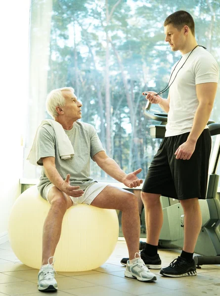Личный тренер объясняет старшему человеку, как делать упражнения на фитнес-мяч — стоковое фото