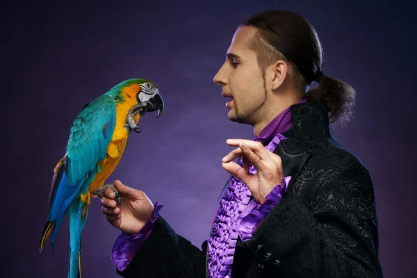 Junge hübsche brünette Zauberer Mann im Bühnenkostüm mit seinem trainierten Papagei — Stockfoto