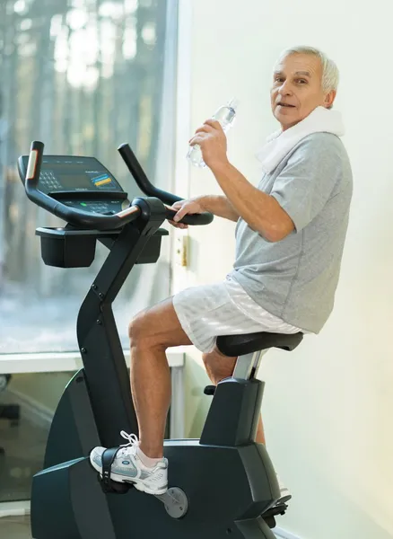 Hombre mayor con botella de agua en una bicicleta en un gimnasio —  Fotos de Stock