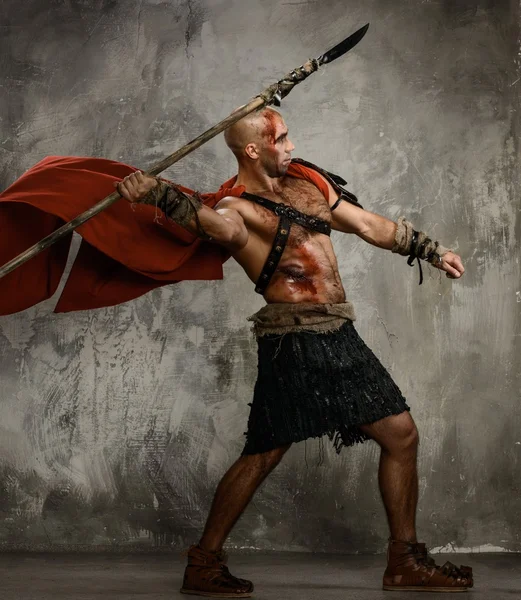 Wounded gladiator in red coat throwing spear — Stock Photo, Image