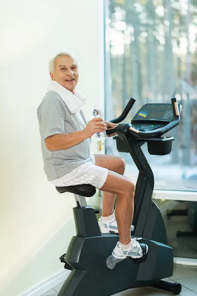 Senior man met fles water op een fiets in een fitnessclub — Stockfoto