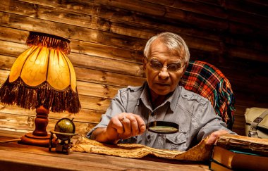 Senior man with magnifier reading vintage book in homely wooden interior  clipart