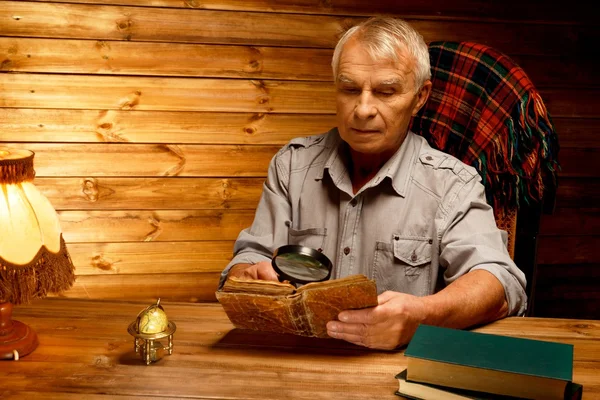 Uomo anziano con lente d'ingrandimento lettura libro d'epoca in interni in legno casalingo — Foto Stock