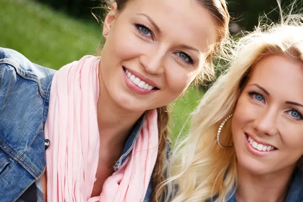 Две красивые улыбающиеся девушки в джинсовых куртках сидят на скамейке в парке — стоковое фото