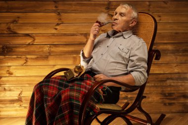 Senior man with smoking pipe sitting on rocking chair in homely wooden interior clipart