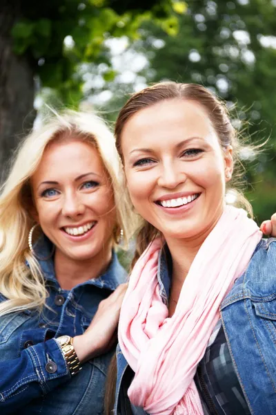 Две красивые улыбающиеся девушки в джинсовых куртках сидят на скамейке в парке — стоковое фото