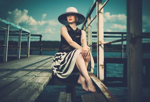 Mulher bonita usando chapéu e cachecol branco sentado no velho cais de madeira — Fotografia de Stock