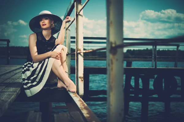 Beautiful woman wearing hat and white scarf sitting on old wooden pier — Stock Photo, Image