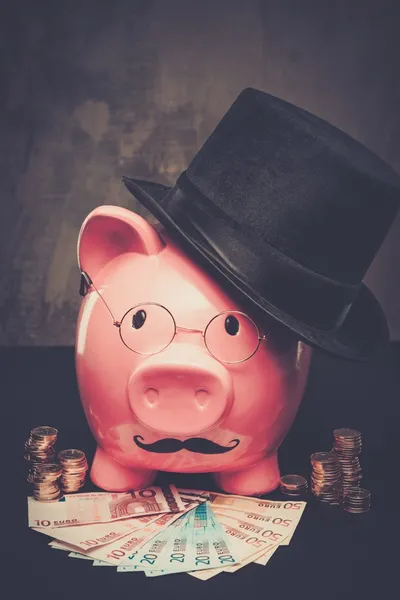 Piggybank em óculos e chapéu com pilha de moedas e notas — Fotografia de Stock