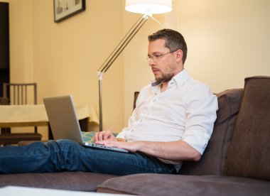 Young man in glasses with laptop on sofa in home interior  clipart