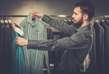 Handsome man with beard choosing shirt in a shop clipart