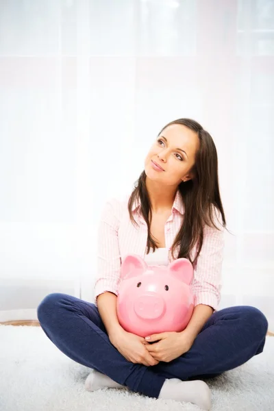 Cheerful young woman sitting on a carpet with piggybank — Stock Photo, Image