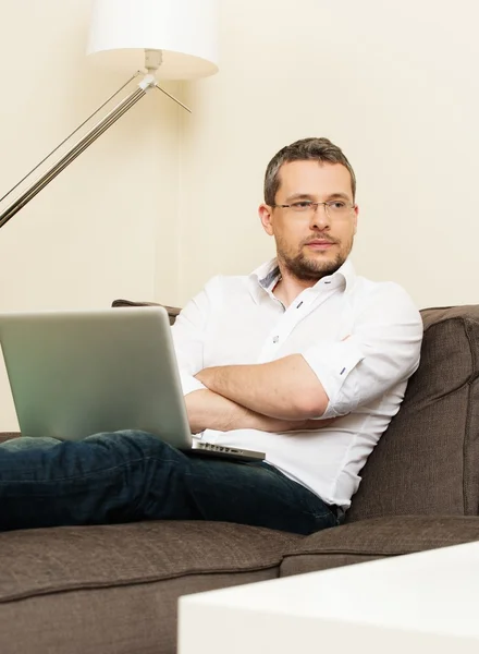 Молодой человек в очках с ноутбуком на диване в интерьере дома — стоковое фото