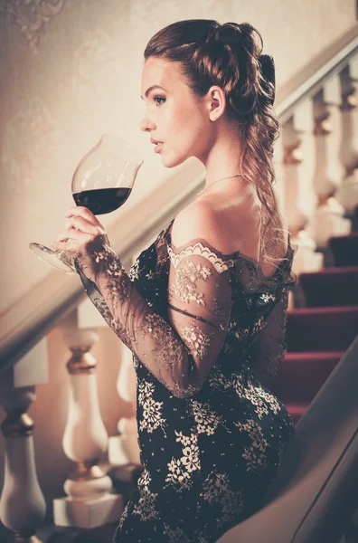 Hermosa mujer joven en vestido de noche largo con copa de vino tinto de pie en una escalera en el interior de lujo —  Fotos de Stock