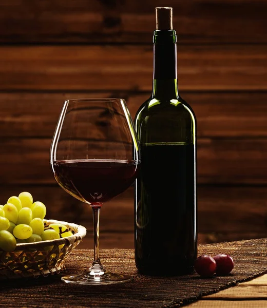 Garrafa de vinho tinto, vidro e uva em cesta no interior de madeira — Fotografia de Stock