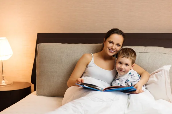 Jovem mãe e seu filho lendo livro em uma cama — Fotografia de Stock