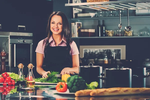 Vrolijke jonge vrouw in schort op moderne keuken snijden groenten — Stockfoto