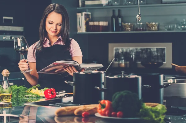 Glad ung kvinna i förkläde på modernt kök med kokbok och glas vin — Stockfoto