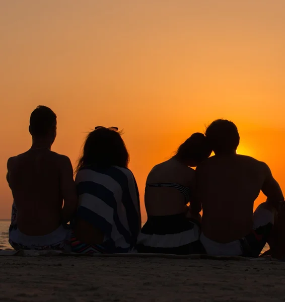 Siluetas una gente joven sentada en una playa mirando el atardecer —  Fotos de Stock